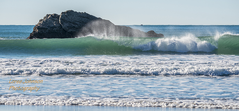 On a beautifully clear, sunny day at the beach, the breeze picks up a bit of the breaking wave, creating a natural water show along the Pacific coast.